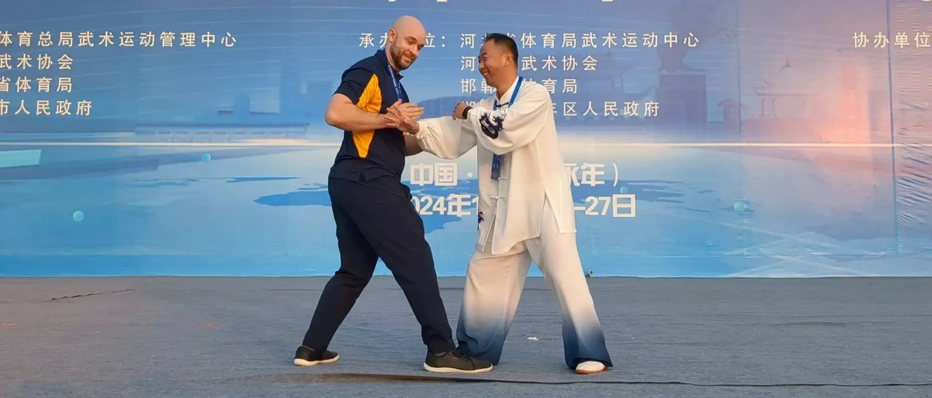 Two adults maintain hand and forearm contact during a standing push-hands demonstration on a conference stage